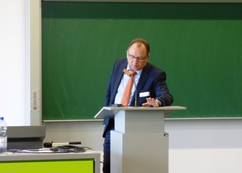 Bild, auf dem Prof. Dr. Gehrmann, Projektleiter des TUD-Sylber, vor einer Tafel an einem Pult mit Mikro steht.