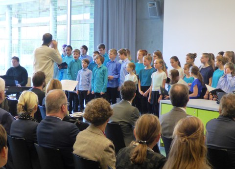 Bild des Chores der Laborschule Dresden vor einem Publikum. Der Chorleiter steht vor den Kindern. Er hält sich eine Stimmgabel an sein Ohr. Ein Mann sitzt neben den Kindern am Klavier, um sie zu begleiten.