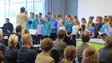 Bild des Chores der Laborschule Dresden vor einem Publikum. Der Chorleiter steht vor den Kindern. Er hält sich eine Stimmgabel an sein Ohr. Ein Mann sitzt neben den Kindern am Klavier, um sie zu begleiten.