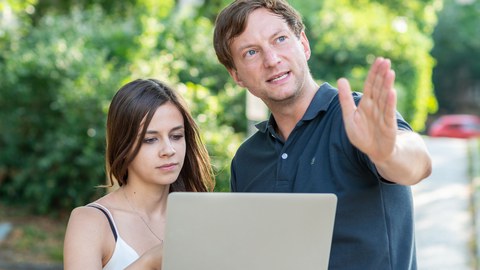 Foto von zwei Personen im Freien, die ein Laptop halten. Der Mann zeigt auf etwas vor ihnen.