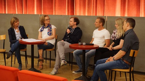 Bild einer Gesprächsrunde von 6 Personen, die im Programmkino Ost um zwei Tische herum sitzen. Ein Mann hat ein Mikrofon in der Hand und redet. Zwei weitere Mikrofone liegen auf dem rechten Tisch.