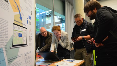 Bild von 4 Personen, die um einen Laptop stehen und in diesen schauen. 2 Männer halten dabei ein Smartphone in ihren Händen.