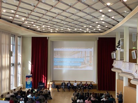 Foto: Blick in einen Festsaal mit Bühne. In Stuhlreihen sitzen zahlreiche Personen.