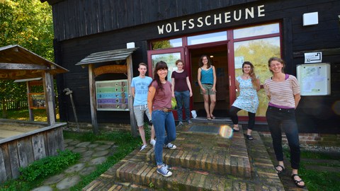 Foto: 6 Personen in Sommerkleidung stehen vor dem Eingang zu einem hölzernen Gebäude. Über der Eingangstür ist die Aufschrift "Wolfsscheune" angebracht.