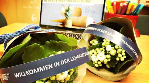 Foto: auf einem Tisch liegen kleine Blumensträuße, auf deren Verpackung "Willkommen in der Lehramtsausbildung" steht. Im Hintergrund ist ein Laptop zu sehen, auf dessen Bildschirm der Text "Willkommen in der Lehramtsausbildung" zu sehen ist.