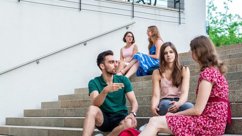 Foto: Fünf Studenten sitzen in lockerer Atmosphäre auf einer Treppe und unterhalten sich.
