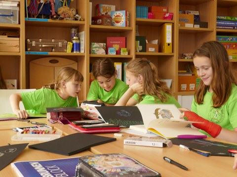 Bild von 4 Mädchen, die um einen Gruppentisch sitzen und Bücher lesen. Im Hintergrund sind Regale mit Büchern und Spielen.
