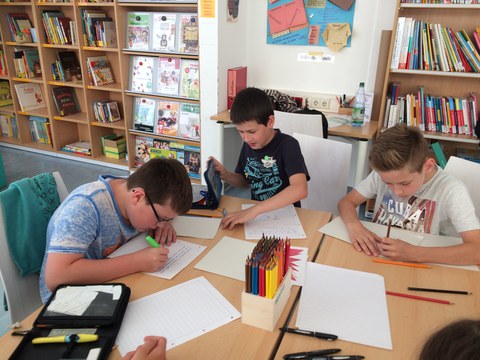 Bild von 3 Kindern, die an einem Gruppentisch sitzen und Arbeitsblätter ausfüllen. Im Hintergrund sind Regale mit Büchern und Zeitschriften.