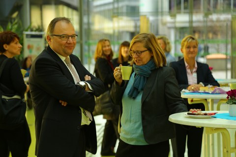 Bild, auf dem ein Mann und eine Frau an einem Stehtisch stehen. Letztere trinkt aus einer Tasse. Im Hintergrund sind weitere Tische und Personen  zu sehen.