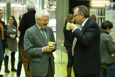 Bild, auf dem zwei Männer einander gegenüber stehen. Beide halten eine Tasse in der Hand. Der linke Mann lacht, der rechte setzt gerade zum trinken an. Im Hintergrund sind weitere Personen und Informationstafeln zu sehen.