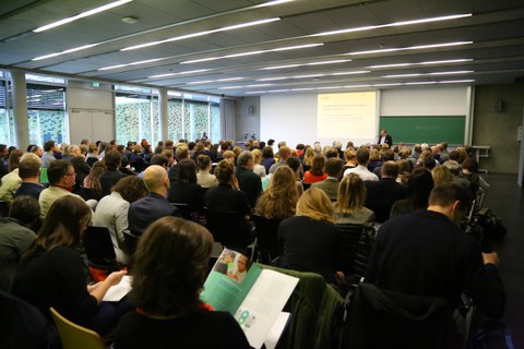 Bild von einem Hörsaal mit Stuhlreihen, der voll besetzt zu sein scheint. Das Publikum hört einer Person zu, die vorne vor einer Tafel an einem Pult steht und augenscheinlich einen Vortrag hält. Links von der Tafel wird etwas an die Wand projiziert.