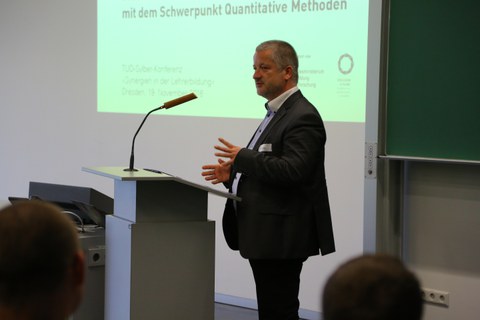 Bild von einem Mann, der vor einer Tafel an einem Pult mit Mikrofon steht und spricht. Links von der Tafel wird eine Präsentation an die Wand projiziert.