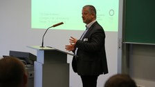 Bild von einem Mann, der vor einer Tafel an einem Pult mit Mikrofon steht und spricht. Links von der Tafel wird eine Präsentation an die Wand projiziert.