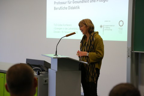 Bild von einer Frau, die vor einer Tafel an einem Pult mit Mikrofon steht und spricht. Links von der Tafel wird eine Präsentation an die Wand projiziert.