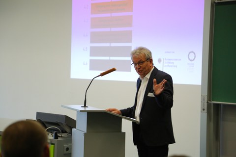 Bild von einem Mann, der vor einer Tafel an einem Pult mit Mikrofon steht und spricht. Links von der Tafel wird eine Präsentation an die Wand projiziert.