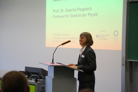 Bild von einer Frau, die vor einer Tafel an einem Pult mit Mikrofon steht und spricht. Links von der Tafel wird eine Präsentation an die Wand projiziert.