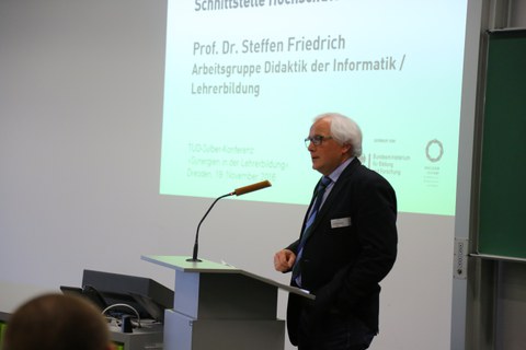 Bild von einem Mann, der vor einer Tafel an einem Pult mit Mikrofon steht und spricht. Links von der Tafel wird eine Präsentation an die Wand projiziert.