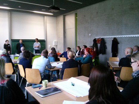 Bild eines Seminarraumes im Hörsaalzentrum der TU Dresden, in dem im Vordergrund mehrere Personen an verschiedenen Gruppentischen sitzen. Im Hintergrund stehen 3 Menschen vor einer Tafel und präsentieren.