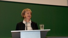 Bild von einer Frau, die vor einer Tafel an einem Pult mit Mikrofon steht und spricht.