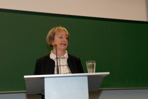 Bild von einer Frau, die vor einer Tafel an einem Pult mit Mikrofon steht und spricht.
