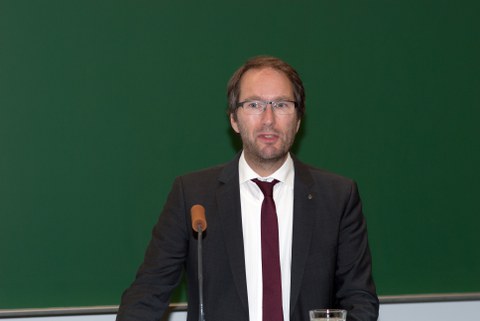 Bild von einem Mann, der vor einer Tafel an einem Pult mit Mikrofon steht und spricht.