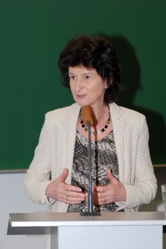 Bild von einer Frau, die vor einer Tafel an einem Pult mit Mikrofon steht und spricht.