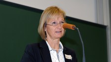 Bild von einer Frau, die vor einer Tafel an einem Pult mit Mikrofon steht und spricht.