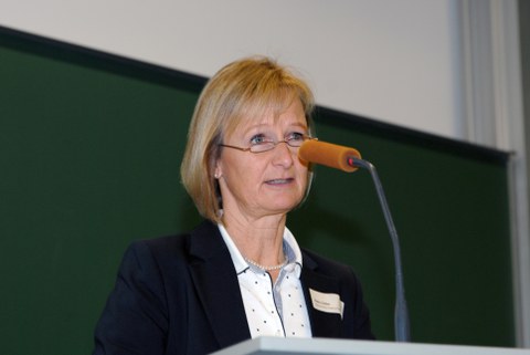 Bild von einer Frau, die vor einer Tafel an einem Pult mit Mikrofon steht und spricht.