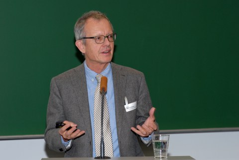 Bild von einem Mann, der vor einer Tafel an einem Pult mit Mikrofon steht und spricht.