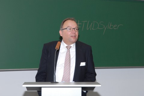 Bild von Prof. Axel Gehrmann. Er steht an einem Pult mit Mikrofon vor einer Tafel und spricht. An der Tafel steht "#TUDSylber".
