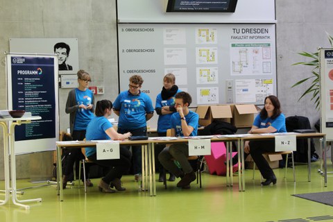 Links im Bild steht ein Flipchart mit einem Tagesprogramm. Daneben sind drei Tische aufgereiht, an denen jeweils ein Schild hängt, mit jeweils einer Person mit blauem T-Shirt dahinter. Drei weitere gleich gekleidete Personen stehen dahinter.