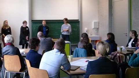 Bild eines Seminarraumes am Hörsaalzentrum der TU Dresden. Im Vordergrund sitzen einige Erwachsene an verschiedenen Gruppentischen. Sie hören den vier Präsentierenden zu, welche nebeneinander vor der Tafel stehen und reden.