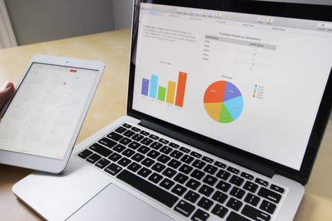 Bild eines Laptops, der offen auf einem Tisch steht. Eine Tabelle und zwei Diagramme sind auf dem Bildschirm zu erkennen. Im linken Teil des Bildes hält eine Hand ein Tablet, auf dem ein Kalender geöffnet ist.