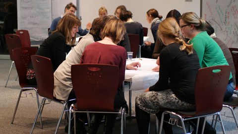 Bild von einem Raum, in dem mehrere Personen um zwei Tische herum verteilt sitzen und miteinander sprechen. Im Hintergrund sind mehrere beschriebene Flipchart-Blätter zu sehen.