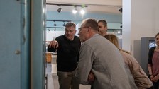 Mann zeigt und erklärt mehreren Personen Ausstellungsstücke, die sich in einem Schrenk befinden.