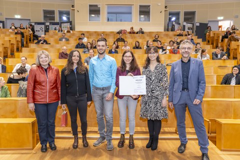 Auf dem Foto sind zwei Männer und vier Frauen abgebildet, die nebeneinander stehen und für eine Preisentgegennahme posieren. IM Hintergrund ist eine Hörsaal mit Holzbestuhlung zu sehen