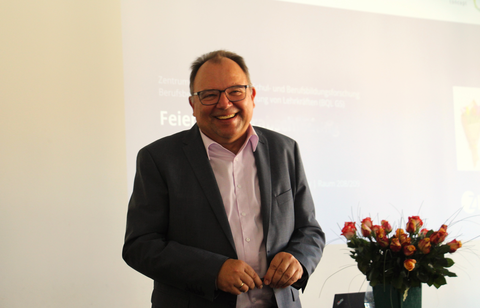 Man sieht Prof. Gehrmann, der die neuen Teilnehmenden in der wissenschaftlichen Ausbildung mit Grußworten und Wünschen fröhlich willkommen heißt. Im Hintergrund sind Blumen und ein Beamer-Bild.
