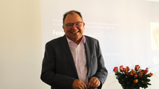 Man sieht Prof. Gehrmann, der die neuen Teilnehmenden in der wissenschaftlichen Ausbildung mit Grußworten und Wünschen fröhlich willkommen heißt. Im Hintergrund sind Blumen und ein Beamer-Bild.