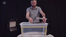 Auf dem Bild erklärt der Dozent den Aufbau für die Videoproduktion anhand einer Trickfilmbox. Neben der Trickfilmbox sind rechts und links Lampen aufgestellt. Auf der Box liegt oben auf dem Deckel ein Tablet für die Videoaufnahme.