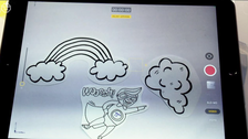 Auf dem Bild sieht man ein Tablet, auf dem gerade eine Szene aus einem Legetechnik-Video abgespielt wird: Man Sieht in der Szene drei Papier-Symbole: Regelwolke, Wolke und einen Comic-Superman, der durch die Luft fliegt.