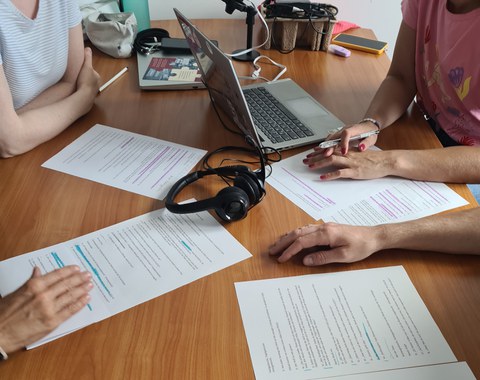 Nahaufnahme eines Tisches mit Notizen, Laptop und Kopfhörern. 4 Personen sitzen an diesem Tisch und arbeiten gemeinsam