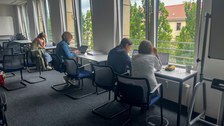Personen sitzen an Tischen am Fenster und arbeiten einzeln an Aufgaben