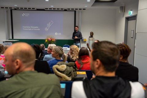 Mann im Hörsaal spricht neben Präsentationsfolie mit dem Titel: "Feierliche übergabe . Transcripts of Records"