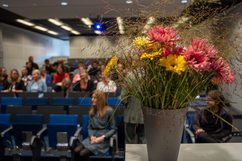 Blumenstrauß auf Tisch, im Hintergrund sitzen Personen im Hörsaal