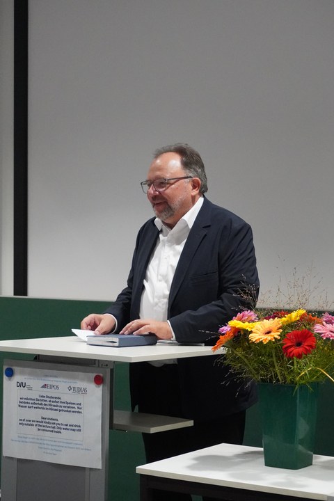 Mann steht an Rednerpult neben Strauß Blumen