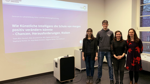 Drei Frauen und ein Mann stehen vor einer Wand, auf der eine Titelseite einer Präsentation projiziert wird. 