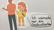 Auf dem Bild, das mit Papierfiguren für die Legetechnik entstanden ist, sieht man ein Mädchen, das zu ihren Papa in einer Sprechblase sagt: "Ich wünsche mir ein Geschwisterchen"