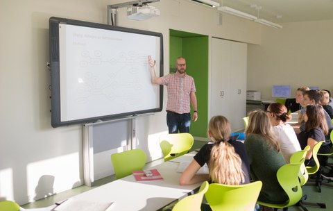 Bild, auf dem ein Lehrer an einer interaktiven Tafel steht und etwas erklärt. Vor ihm sitzen mehrere Schüler:Innen in einer Reihe und hören zu.