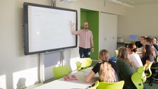 Bild, auf dem ein Lehrer an einer interaktiven Tafel steht und etwas erklärt. Vor ihm sitzen mehrere Schüler:Innen in einer Reihe und hören zu.