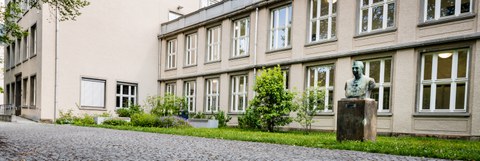 Bild einer Hausansicht des Physikgebäudes "Recknagel-Bau" an der TU Dresden. Zu sehen ist das Gebäude, davor ein Weg mit Grünfläche und einer Büste.